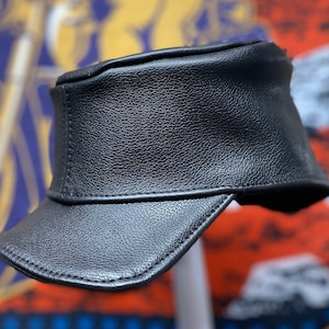 May include: A black leather cap with a short brim. The cap has a structured design and a textured surface. The background is blurred, but hints at a motorcycle-themed design.
