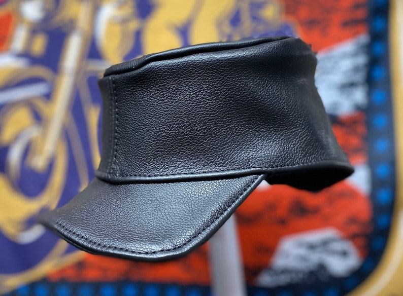May include: A black leather cap with a short brim. The cap has a structured design and a textured surface. The background is blurred, but hints at a motorcycle-themed design.
