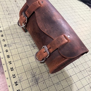 Pode incluir: Uma bolsa de sela de motocicleta de couro marrom com duas fivelas e um anel de metal. A bolsa est&aacute; desgastada e tem um aspecto desgastado.