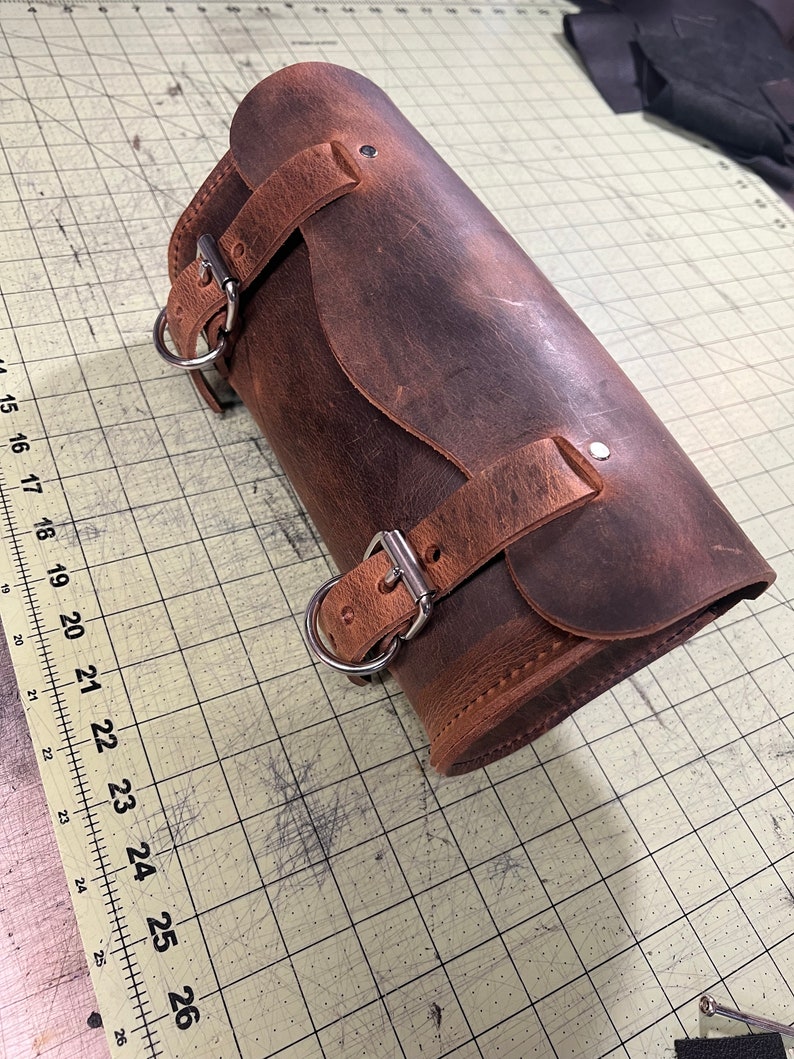 Pode incluir: Uma bolsa de sela de motocicleta de couro marrom com duas fivelas e um anel de metal. A bolsa est&aacute; desgastada e tem um aspecto desgastado.