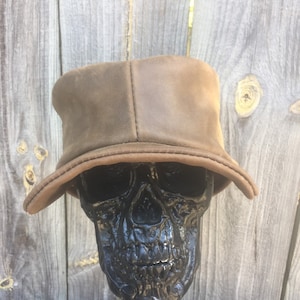 May include: A brown leather bucket hat with a slightly curved brim. The hat is displayed on a black skull sculpture. The hat has a soft, worn appearance, and the brim is a lighter shade of brown.