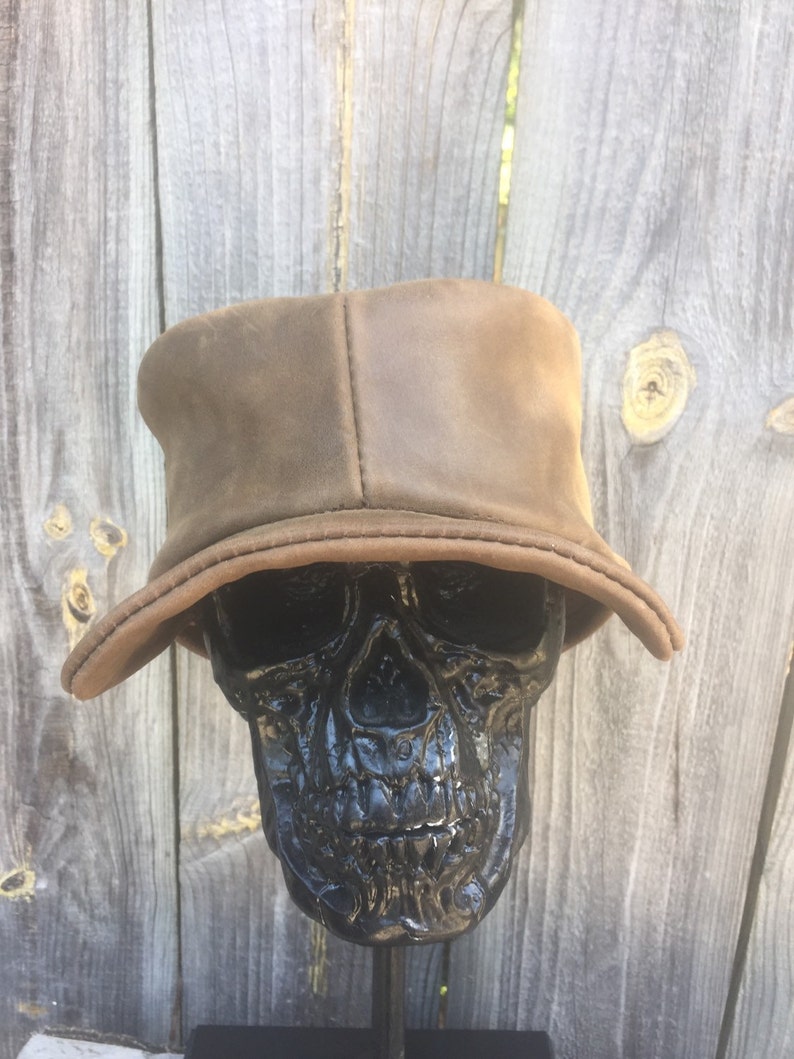 May include: A brown leather bucket hat with a slightly curved brim. The hat is displayed on a black skull sculpture. The hat has a soft, worn appearance, and the brim is a lighter shade of brown.