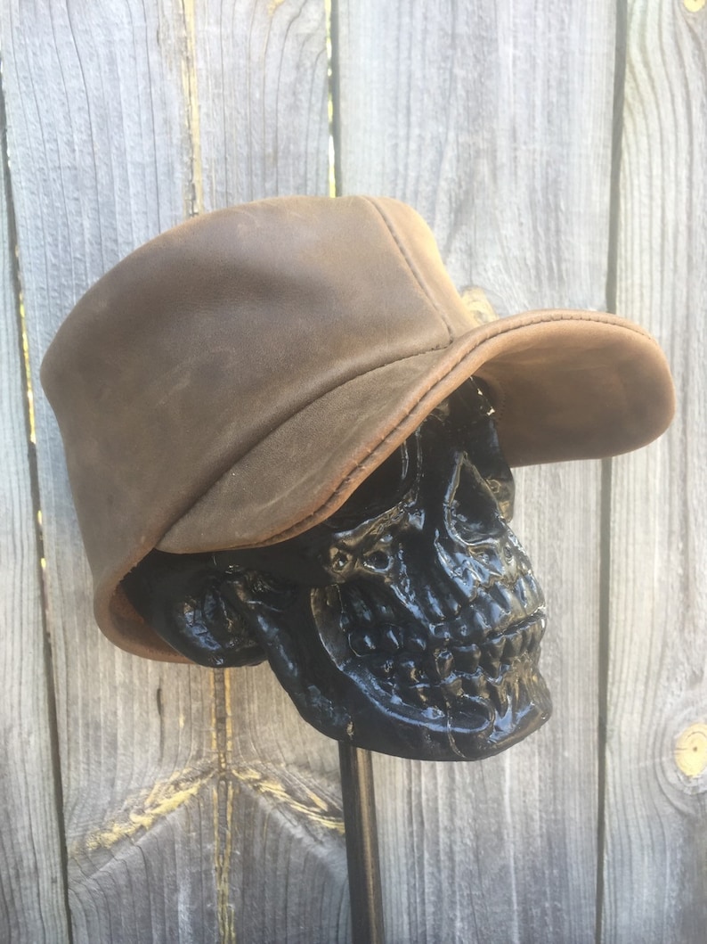 May include: A brown leather hat with a curved brim sits atop a black skull sculpture. The hat has a distressed appearance, with visible stitching and a slightly worn texture. The skull is glossy and detailed.