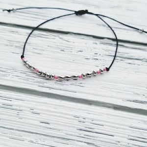 Puede incluir: Un brazalete sencillo con un cord&oacute;n negro y cuentas plateadas y rosadas.