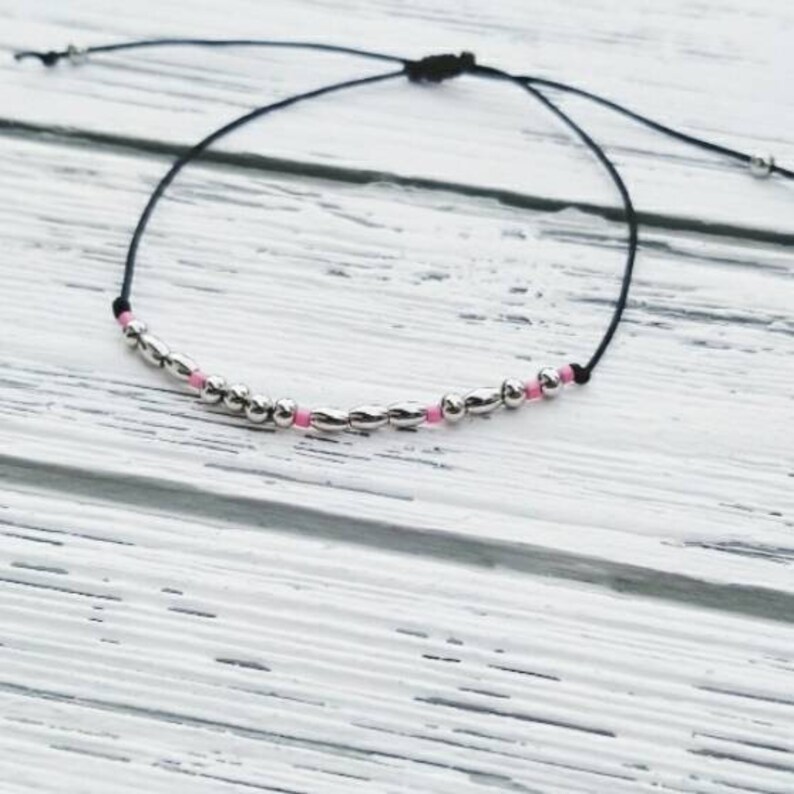 Puede incluir: Un brazalete sencillo con un cord&oacute;n negro y cuentas plateadas y rosadas.