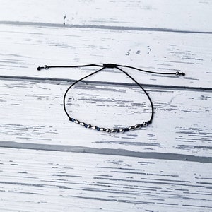 May include: A black cord bracelet featuring a series of small, metallic silver-toned beads interspersed with tiny, dark blue cylindrical beads. The bracelet includes an adjustable sliding knot closure with a small silver bead at each cord end.
