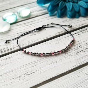 May include: A simple black string bracelet with silver and pink beads. The bracelet is adjustable and can be worn on the wrist or ankle.
