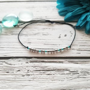 May include: A black cord bracelet featuring a series of metallic silver beads and small turquoise blue beads arranged in a linear pattern. The bracelet has a thin, adjustable black string design with silver end beads on each side of the cord.