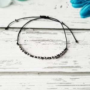 May include: A delicate adjustable bracelet featuring a thin black cord strung with a series of small, polished silver-toned metallic beads. The design includes two additional silver beads on the cord ends, which serve as a sliding closure mechanism.
