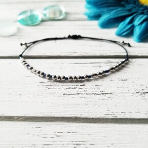May include: A silver and blue beaded bracelet with a black cord. The bracelet is on a white wooden surface.