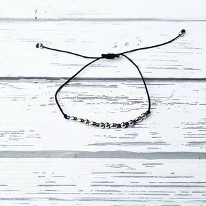 May include: A black cord bracelet featuring a series of small, polished silver-toned metallic beads interspersed with tiny dark blue accent beads. The bracelet includes an adjustable sliding knot closure with two small silver-toned beads at the ends.