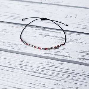 May include: A delicate bracelet featuring a thin black cord strung with a sequence of small, metallic silver-toned beads and tiny red seed beads. The adjustable cord ends in two small silver-toned beads, creating a simple and minimalist design.