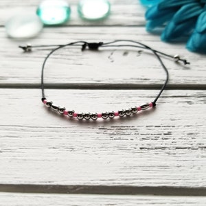 May include: A black string bracelet with silver and pink beads. The bracelet is adjustable and can be worn on the wrist or ankle.