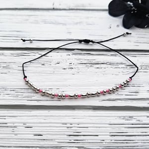 May include: A black cord bracelet featuring a series of alternating silver-toned metallic beads and small pink beads. The bracelet includes an adjustable sliding knot closure with two small silver-toned beads at the ends of the black cord strings.