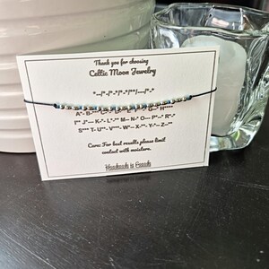 May include: A handmade bracelet with silver and blue beads, strung on a black cord, is displayed on a card that reads "Thank you for choosing Celtic Moon Jewelry." The card also includes a Morse code key and the text "Handmade in Canada."