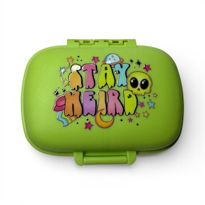 May include: A lime green, rectangular, hinged container with the words "STAY WEIRD" in colorful, bubble-style letters. The design includes a cartoon skull, stars, a crescent moon, and a planet.