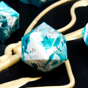 Peut inclure: Ensemble de dés polyédriques avec un motif marbré turquoise, blanc et argenté. Les dés ont une finition brillante et sont posés sur un fond noir avec un élément décoratif doré. Idéal pour les jeux de société.