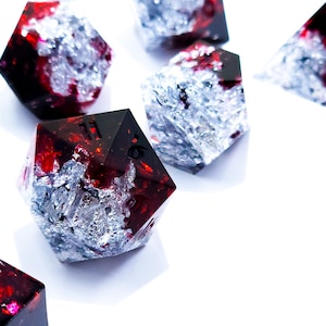 May include: Set of polyhedral dice with a striking design. The dice feature a combination of deep red and silver foil embedded in a clear resin, creating a unique visual effect. The dice are scattered on a white surface.