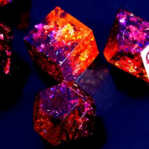 May include: A set of six-sided dice with a red and purple iridescent finish. The dice are made of resin and have a glittery appearance. The numbers on the dice are visible.