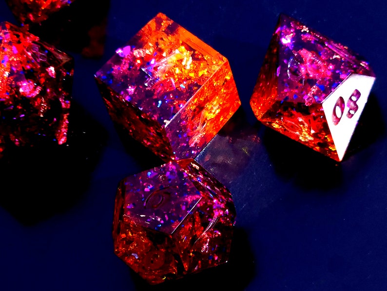 May include: A set of six-sided dice with a red and purple iridescent finish. The dice are made of resin and have a glittery appearance. The numbers on the dice are visible.