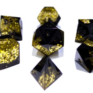 May include: A set of seven polyhedral dice in various shapes, including a d20, d6, and d4. The dice are black with gold glitter embedded within the resin. The dice are arranged on a white surface.