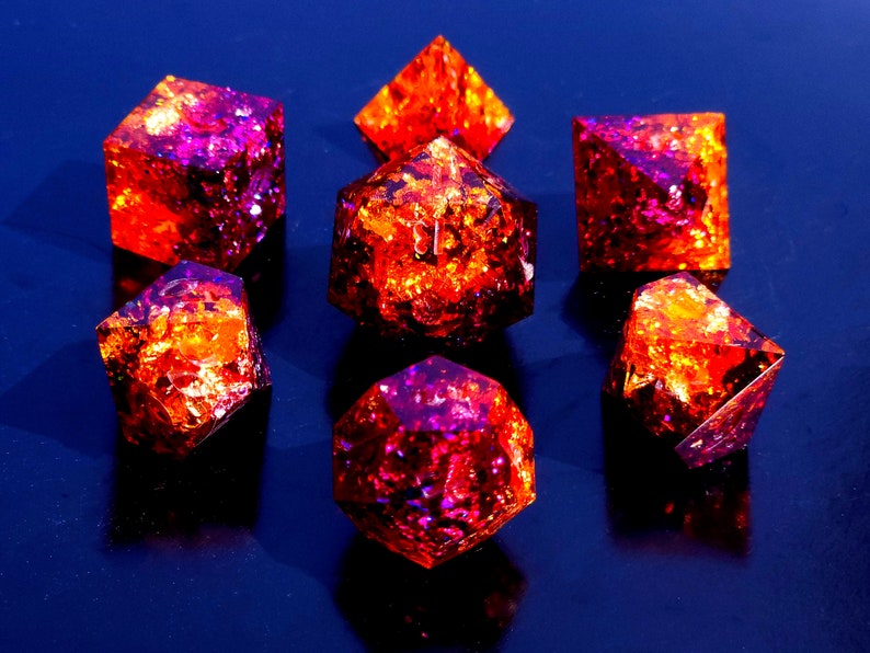 May include: Set of seven iridescent dice with a red and purple color scheme. The dice are made of resin and have a glittery finish.