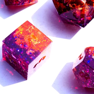 May include: Four iridescent dice with a gradient of purple and orange glitter. The dice are made of resin and have a variety of numbers on their faces.