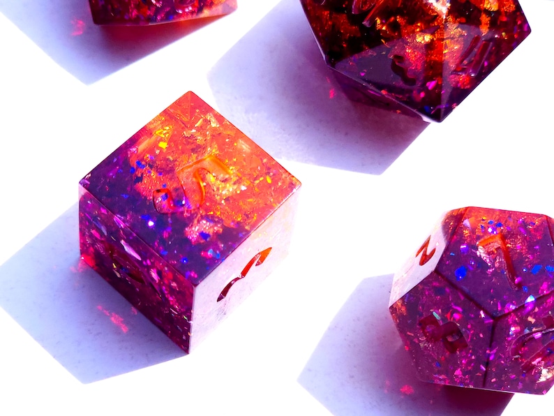 May include: Four iridescent dice with a gradient of purple and orange glitter. The dice are made of resin and have a variety of numbers on their faces.