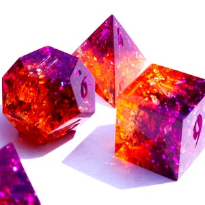 May include: A set of six-sided dice with a purple and orange gradient and gold glitter embedded in the resin. The dice are in various shapes, including a pyramid, a cube, and a dodecahedron.