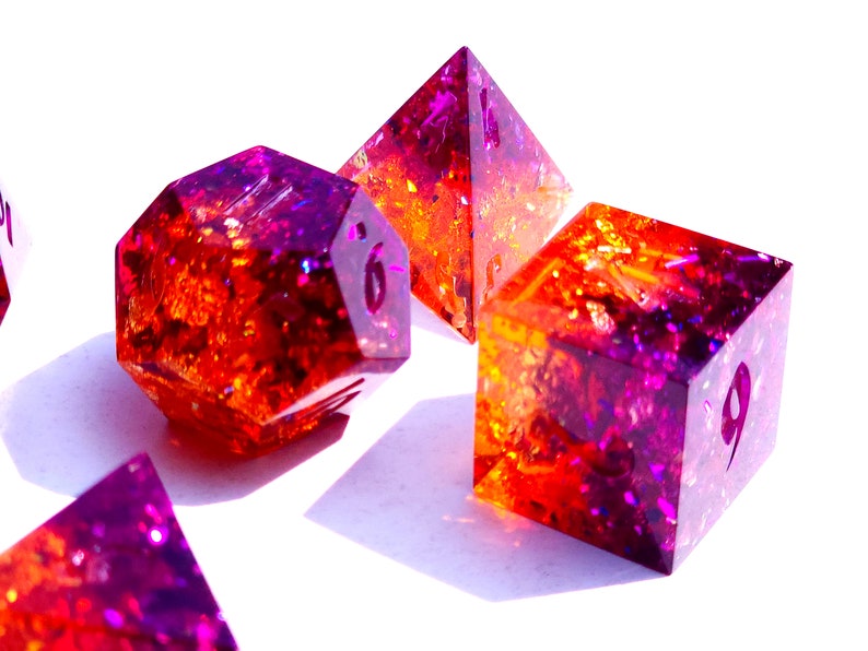 May include: A set of six-sided dice with a purple and orange gradient and gold glitter embedded in the resin. The dice are in various shapes, including a pyramid, a cube, and a dodecahedron.