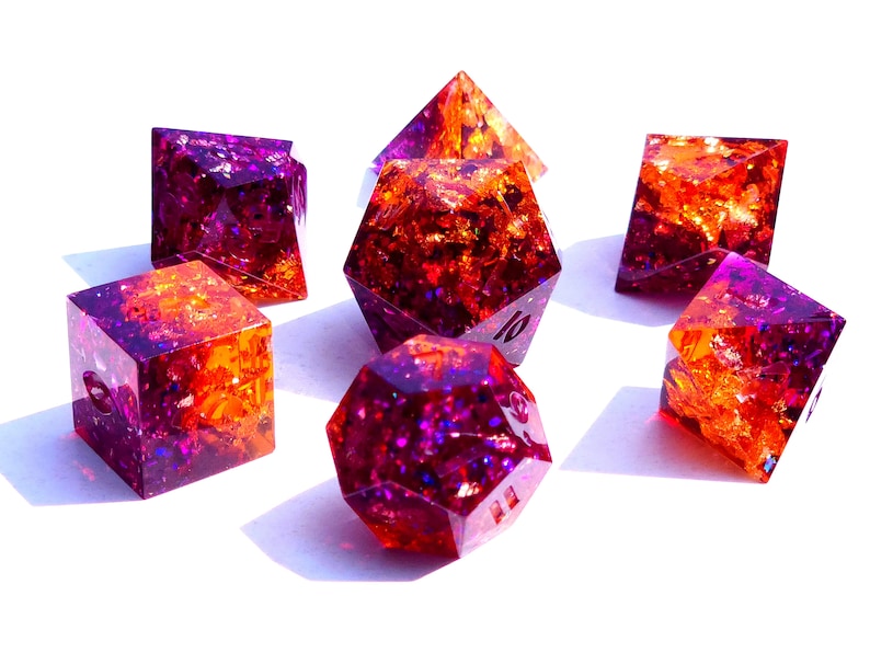 May include: Set of seven iridescent dice with a purple and orange color scheme. The dice are made of resin and have a glittery finish.