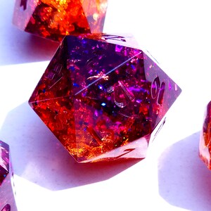 May include: Close-up of four iridescent dice with a purple and orange glitter effect. The dice are made of resin and have a clear, glossy finish.