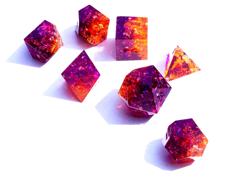 May include: Set of seven iridescent dice with a purple and orange color gradient. The dice are made of resin and have a glittery finish.
