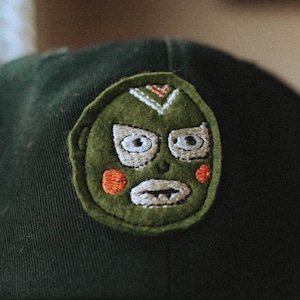 May include: A dark green baseball cap with a green, white, and orange embroidered patch of a luchador mask.
