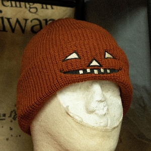 Puede incluir: Un gorro de punto marrón con una cara de calabaza bordada en la parte delantera. El gorro está en una cabeza de maniquí blanca.