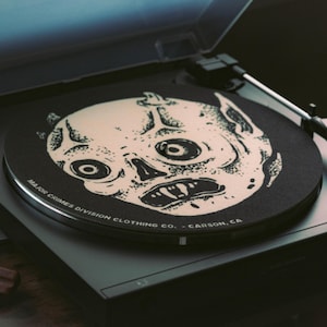 May include: A black turntable with a vinyl record featuring a cream-colored, stylized skull design. The record player has a clear cover and the text "MAJOR CRIMES DIVISION CLOTHING CO. - CARSON, CA" is printed on the record.