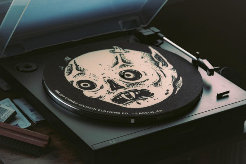 May include: A black turntable with a vinyl record featuring a cream-colored, stylized skull design. The record player has a clear cover and the text "MAJOR CRIMES DIVISION CLOTHING CO. - CARSON, CA" is printed on the record.