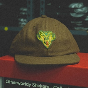 May include: A brown felt baseball cap with a green and yellow embroidered patch of a horned creature with a tongue sticking out. The text "Otherworldy Stickers - Collect them all" is printed on a red box below the cap. The text "MAJOR CRIMES DIVISION CLOTHING CO. - CARSON, CALIFORNIA" is printed below the first line of text. The text "HALLOWEEN", "LOGO COLLECTION", and "SCI-FI" are printed on three separate boxes below the first two lines of text.