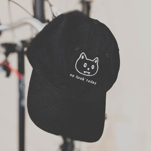 Puede incluir: Gorra de béisbol negra con un rostro de gato bordado en blanco y el texto "no luck today".