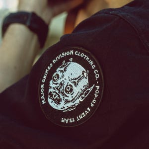 May include: Black jacket with a circular patch featuring a detailed, stylized skull graphic. The patch reads "Major Crimes Division Clothing Co. - Pop-Up Event Team" in a circular design. The image is a close-up of the jacket's shoulder.