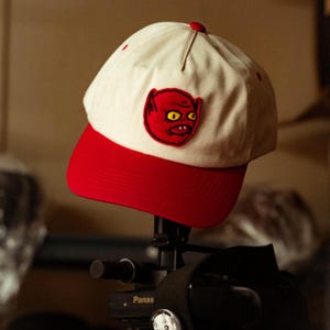 Peut inclure: Casquette de baseball avec une couronne crème et une visière rouge. La casquette présente un écusson rouge brodé d'un diable de dessin animé aux yeux jaunes. La casquette est présentée sur un appareil photo noir.