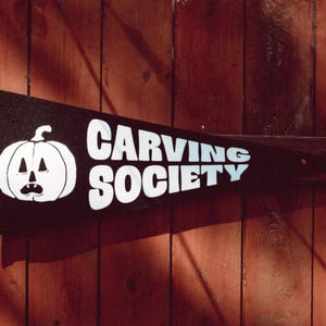 May include: A black pennant with white text that reads "CARVING SOCIETY". A white cartoon pumpkin with a face is on the left side of the pennant.