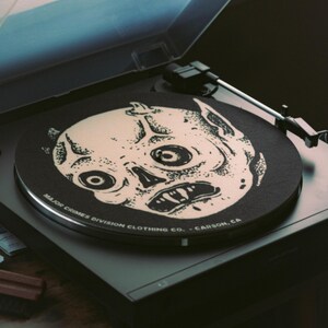 May include: A black record player with a vinyl record featuring a white illustration of a skull-like face. The record player has a clear plastic cover and the text "MAJOR CRIMES DIVISION CLOTHING CO. - CARSON, CA" is printed on the record.