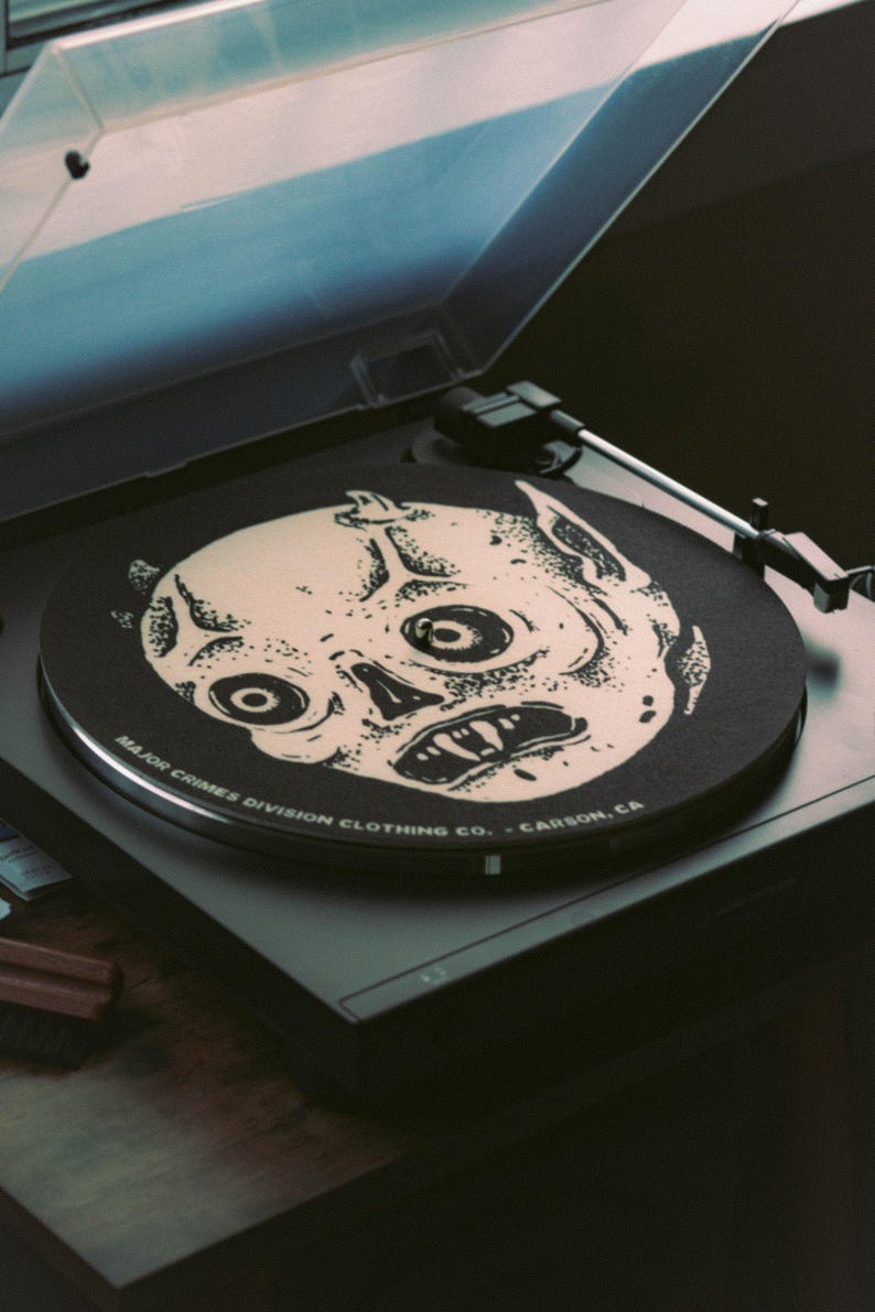 May include: A black record player with a vinyl record featuring a white illustration of a skull-like face. The record player has a clear plastic cover and the text "MAJOR CRIMES DIVISION CLOTHING CO. - CARSON, CA" is printed on the record.