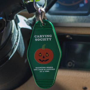 May include: Green keychain tag with the words "CARVING SOCIETY" and a jack-o'-lantern graphic. Below the graphic, the text reads "HAUNTING HOMES ONE JACK-O-LANTERN AT A TIME". The keychain is attached to a set of keys.