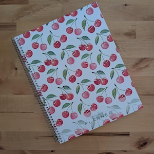 May include: A spiral-bound planner with a white cover featuring a repeating pattern of red cherries and green leaves. The bottom right corner of the cover displays the text Yours Truly PLANNER in a simple, dark, sans-serif font.