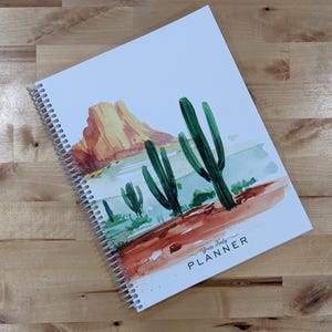 May include: A spiral-bound planner with a white cover featuring a watercolor illustration of a desert landscape with green cacti and a large orange rock formation. The text "Yours Truly PLANNER" is printed in black in the bottom right corner.