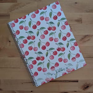 May include: A spiral-bound planner with a white cover featuring a repeating pattern of red cherries and green leaves. The bottom right corner of the cover displays the text Yours Truly PLANNER in a simple, dark, sans-serif font.