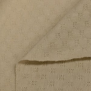 May include: A close-up of a beige knitted fabric with a textured pattern. The fabric is folded in half, showing the pattern on both sides.