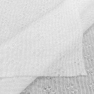 May include: White knitted fabric with a delicate openwork pattern. The fabric is soft and lightweight, making it ideal for a variety of projects.